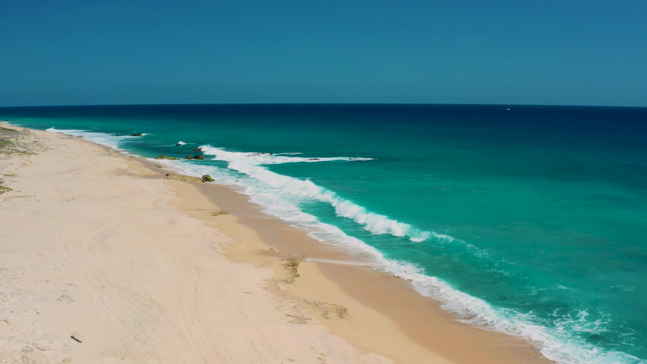 High aerial frame over El Cardoncito, Baja California Sur, with turquoise ocean on the right, pale sand on the left, white surf crossing the shoreline, and no visible people.