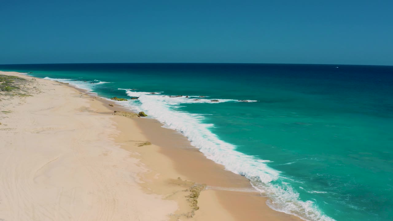 A high coastal view over El Cardoncito shows the shoreline sliding diagonally from lower left to upper center, with whitewater against sand and no visible people.