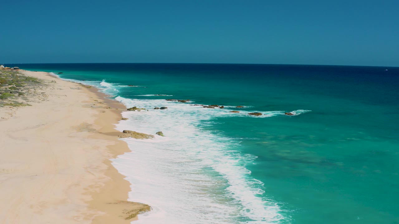 Opening aerial frame over El Cardoncito beach with pale sand on the left, turquoise water on the right, white surf crossing rocks, and no visible people.