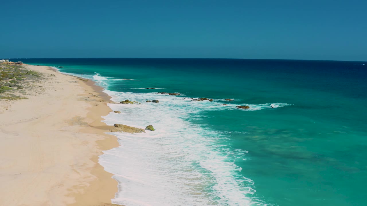 Aerial view at El Cardoncito with the beach tapering into the distance, whitewater curling around scattered rocks, deep blue water near the horizon, and no visible people.