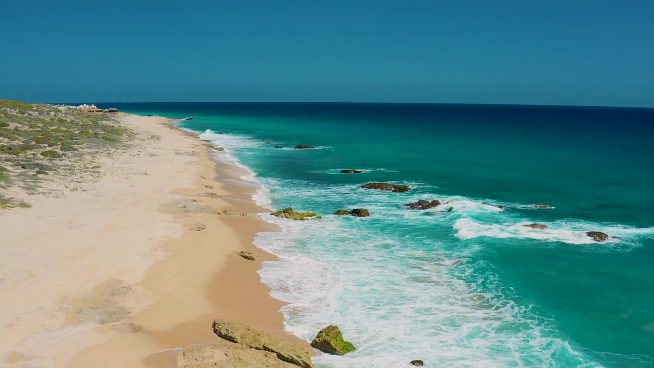 Elevated view along El Cardoncito beach with white surf washing onto the sand, dark rocks offshore, low vegetation on the dune edge, and no visible people.