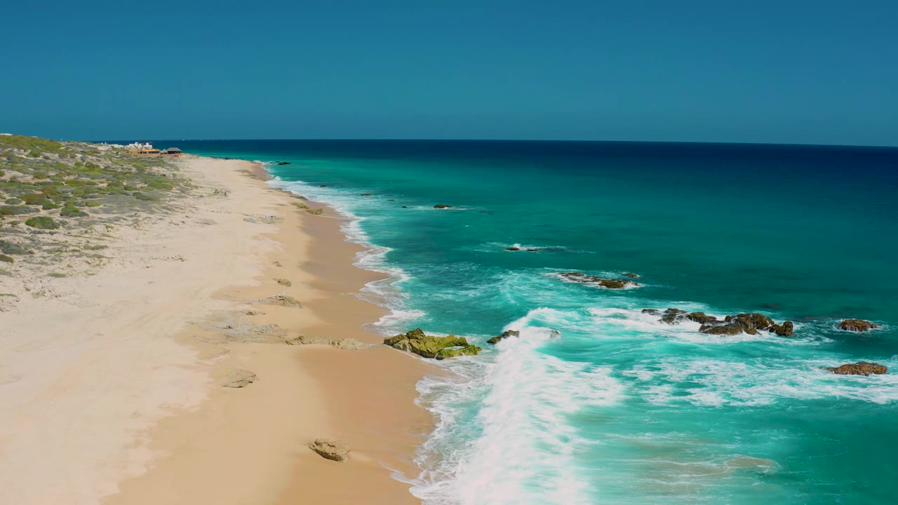 Aerial view of the El Cardoncito beach one second in, with the shoreline running diagonally, white foam spreading near midframe rocks, low vegetation inland, and no visible people.