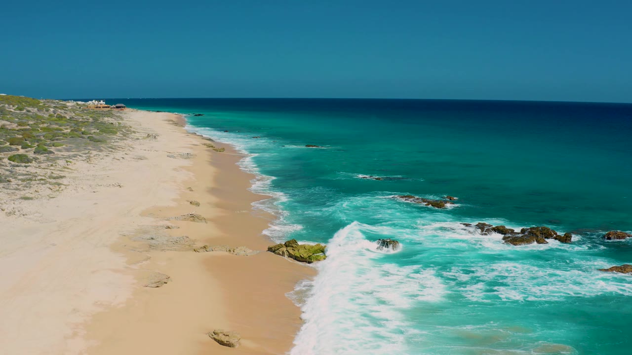 The two-second frame keeps El Cardoncito sand on the left and clear water on the right, with a broad white wave curling around exposed rocks and no visible people.