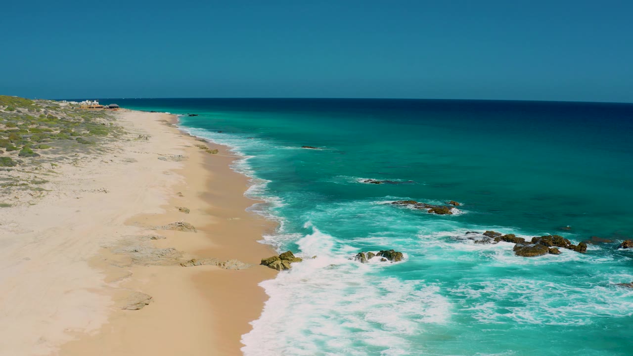 Mid-clip aerial frame over El Cardoncito showing a larger foam band washing toward the sandy beach, dark rocks near the surf edge, open water beyond, and no visible people.