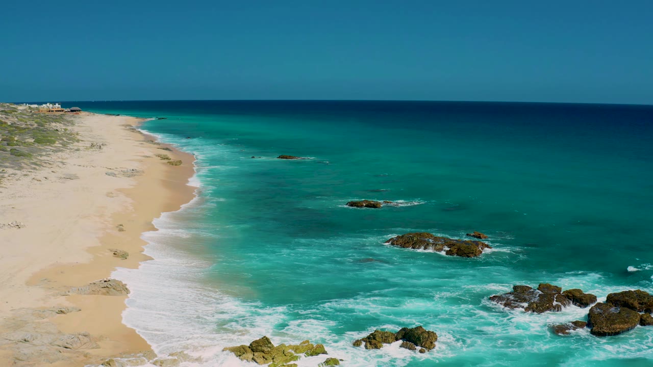 El Cardoncito shoreline seen from above, with a longer sweep of sand at left, white waves curling toward shore, offshore rocks in turquoise water, and no visible people.