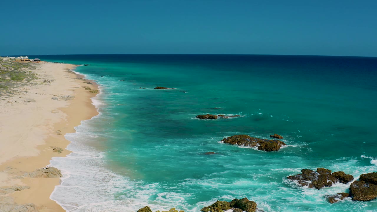 Aerial coastal frame at El Cardoncito showing empty sand along the left border, layered turquoise-to-blue water, several exposed rocks, white foam, and no visible people.
