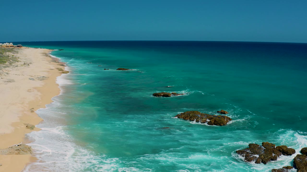 High oblique view of El Cardoncito beach with pale sand at left, a central dark rock surrounded by foam, deeper teal water beyond, and no visible people.
