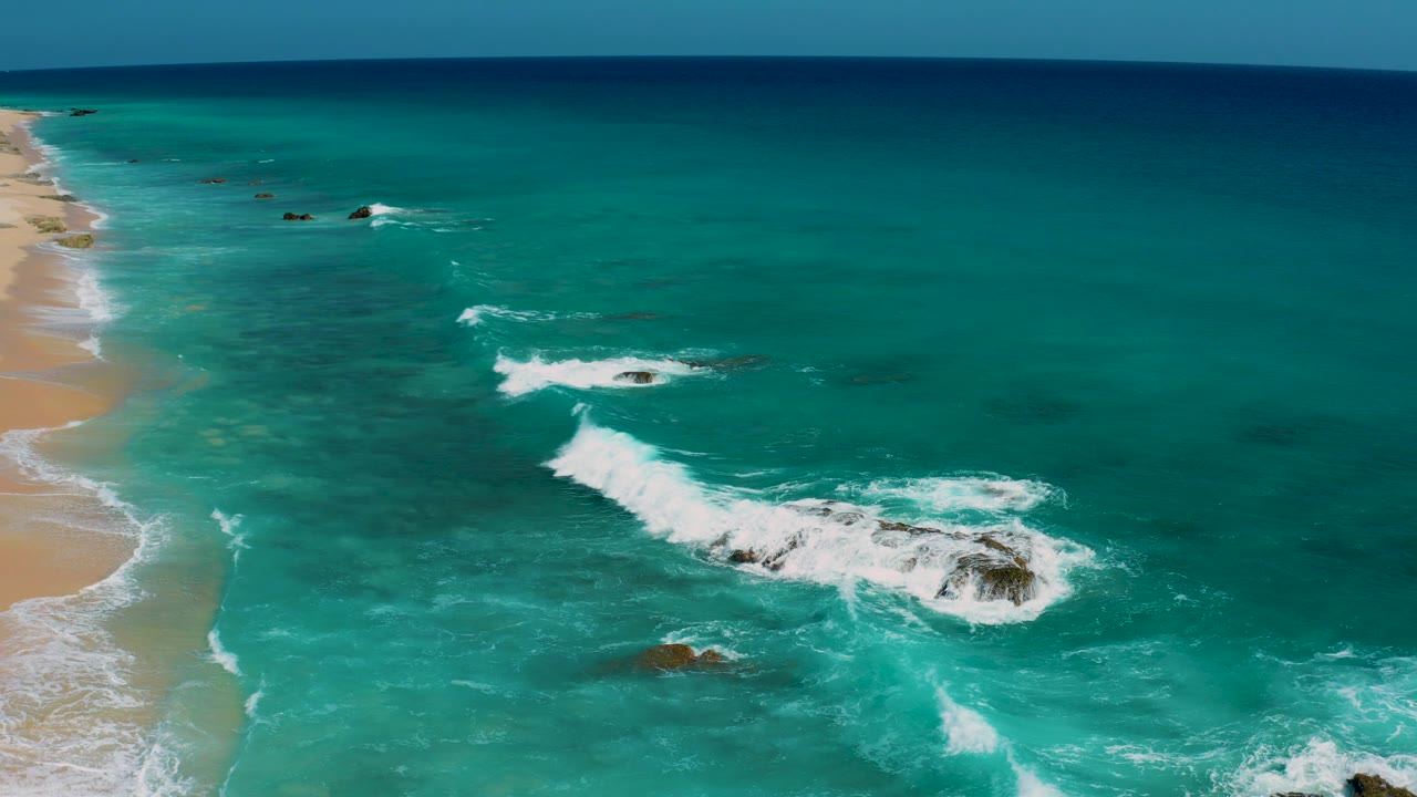 Aerial view of turquoise water, sandy shoreline, and rocks under breaking waves.