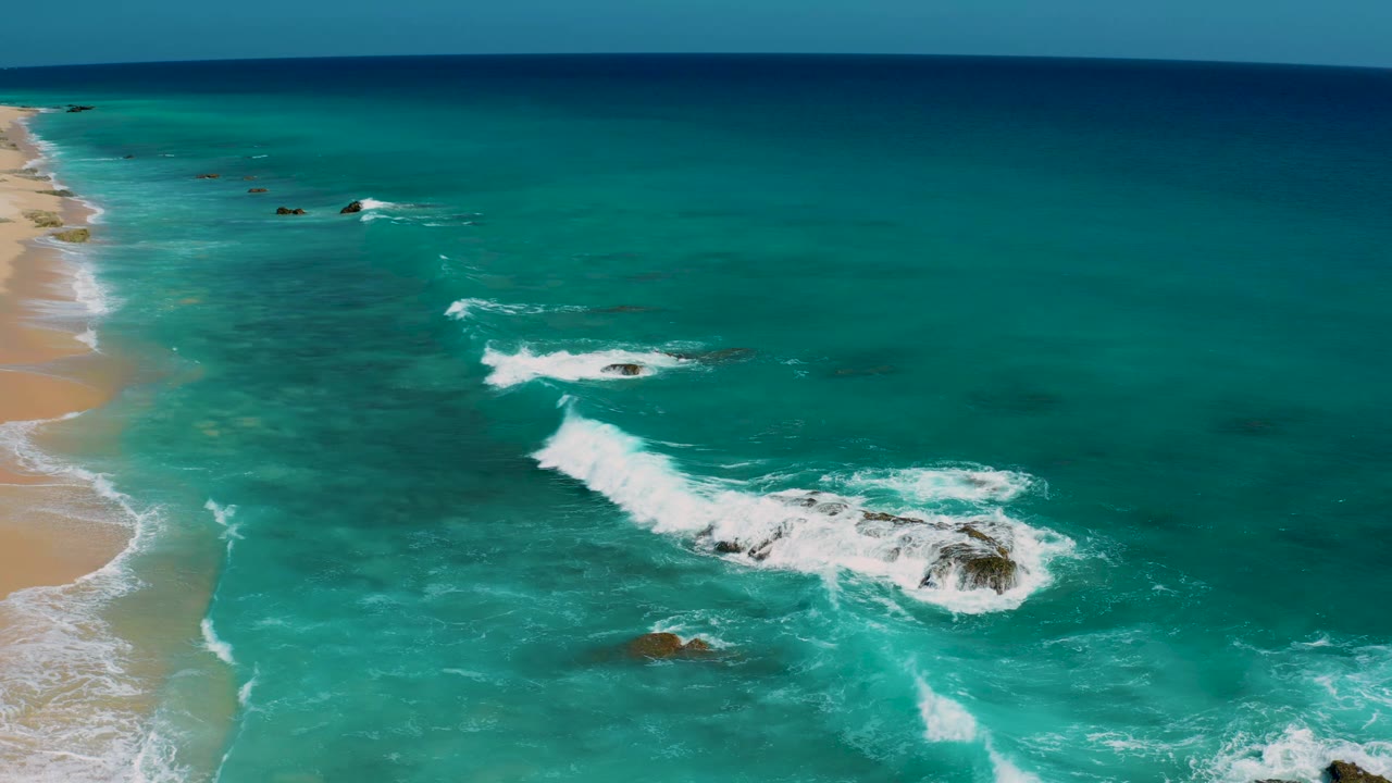 Aerial view at El Cardoncito with the sandy beach along the left side, reef rocks below the surface, white foam curling around a larger rock cluster, and no visible people.