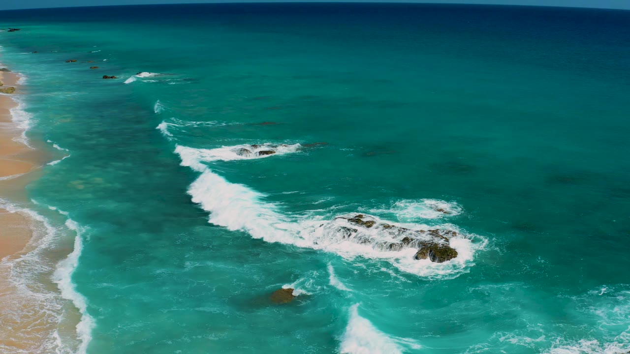 Aerial frame from El Cardoncito showing a diagonal band of white surf over dark rocks, open teal water to the right, sandy shoreline at left, and no visible people.