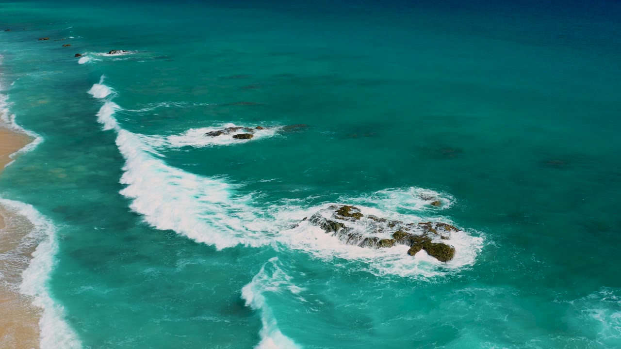 Aerial frame over El Cardoncito with foaming waves spreading from the beach side toward several dark rock outcrops in blue-green water, with no visible people.