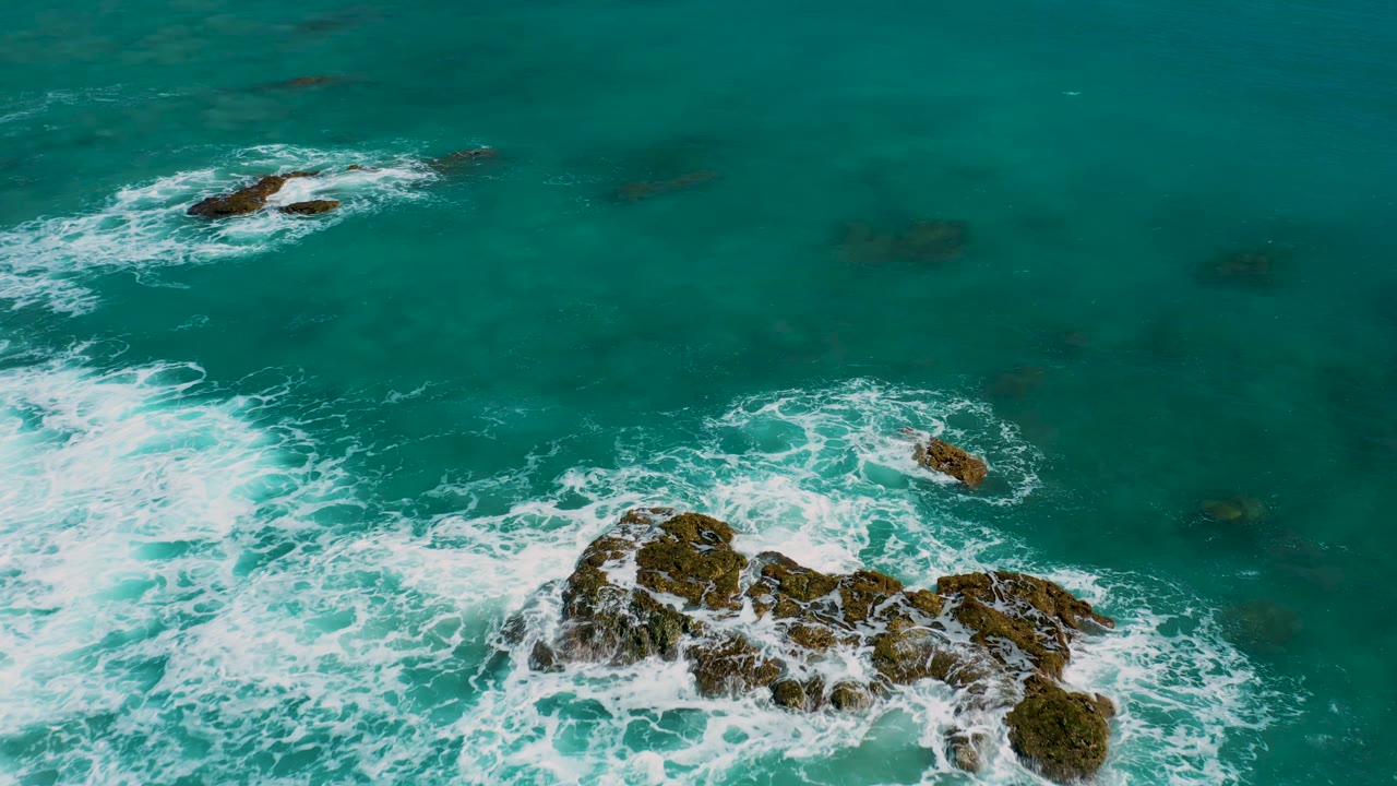 Aerial view in El Cardoncito with the main rock cluster lower in frame, a smaller rock to the right, another outcrop near the top, turquoise-green water, and no visible people.
