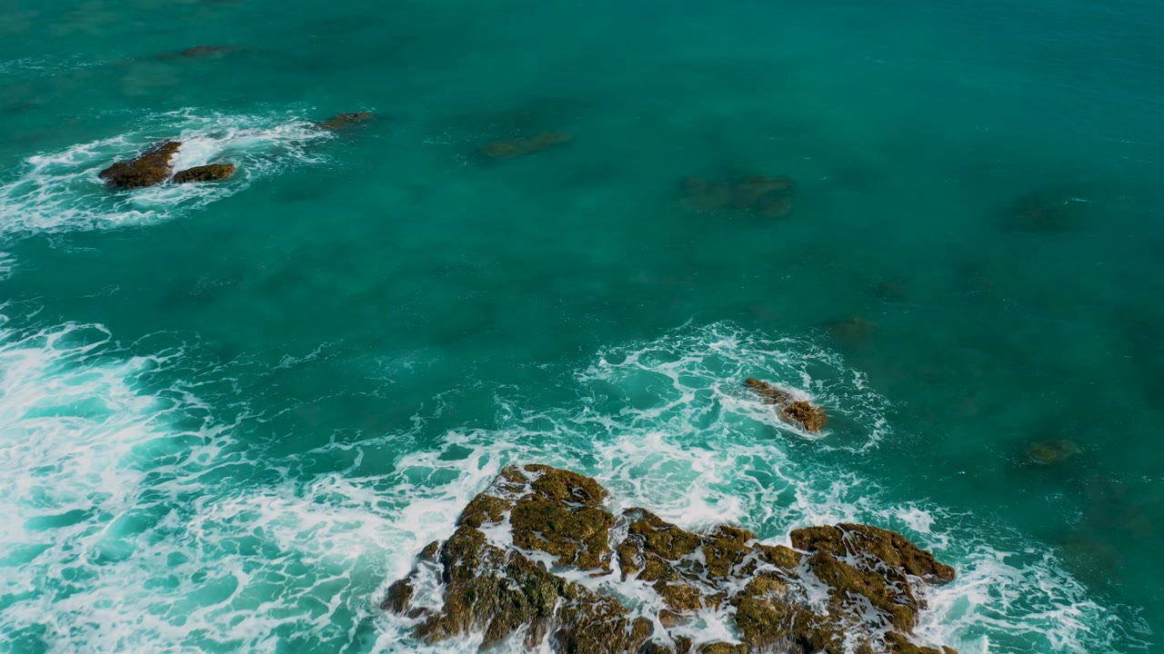 Aerial frame from El Cardoncito showing foamy water curling around a large rock formation near the lower right, smaller rocks above, open green water, and no visible people.
