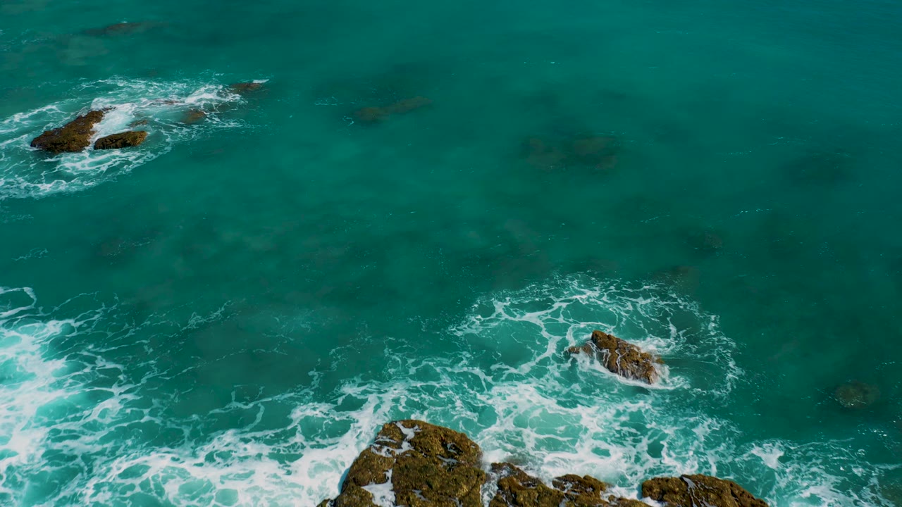 Aerial shot over El Cardoncito water with a large rock cluster partly cropped at the bottom, a smaller foam-ringed rock near center, open green sea, and no visible people.