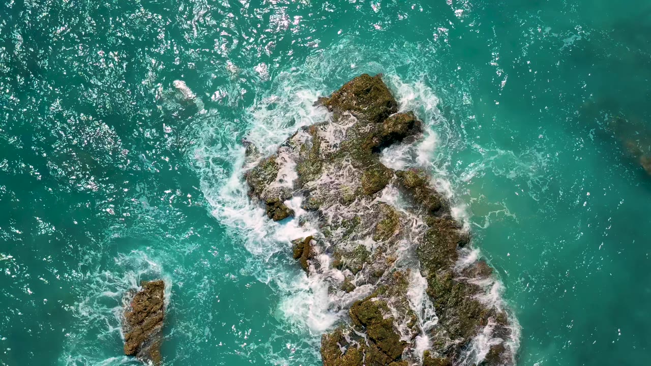 Turquoise sea surrounds the main rocky outcrop and a smaller left-side rock at El Cardoncito, with bright foam collecting along the upper rocks and no visible people.