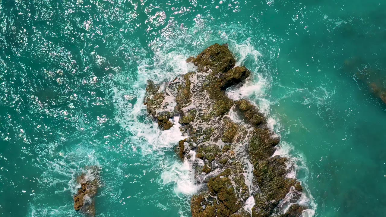 An overhead frame shows El Cardoncito's algae-covered rock mass lower in the composition while foam gathers on the left edge and darker underwater patches show through, with no visible people.