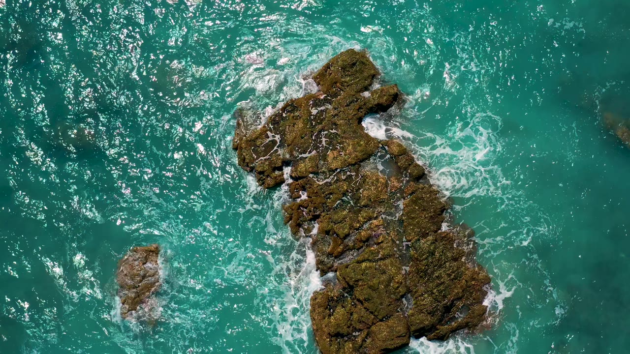 Overhead frame at El Cardoncito, Baja California Sur, with a large dark rock and smaller outcrop surrounded by turquoise water and white foam, no visible people.