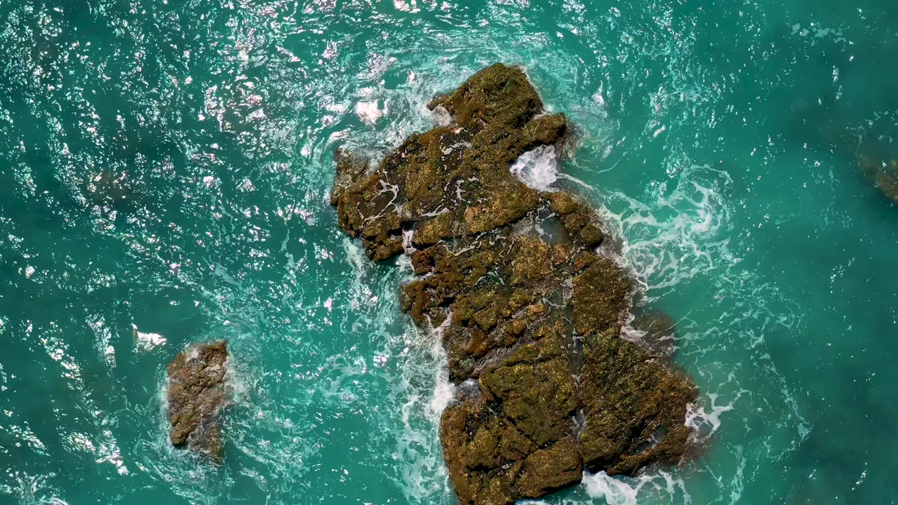 At 0:02, the El Cardoncito rock formation is framed slightly right of center with open teal water around it and a small rock lower left, no visible people.