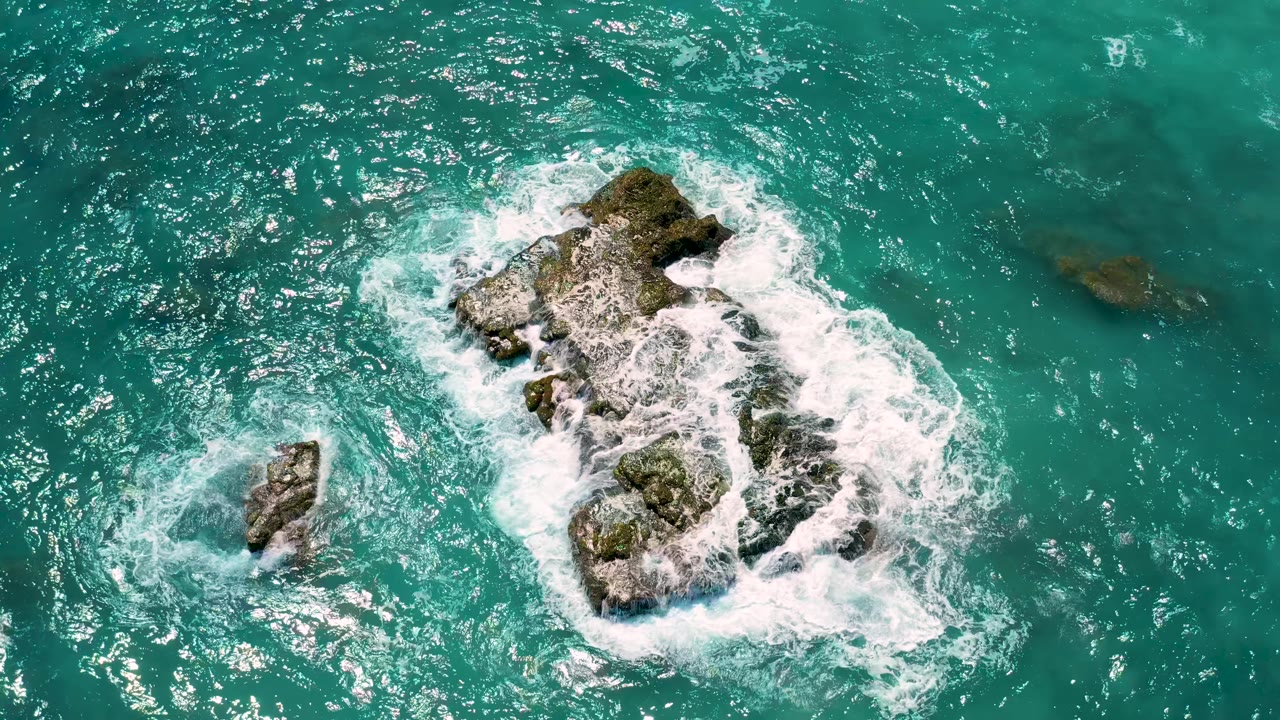 Aerial frame from El Cardoncito, Baja California Sur with the main rock slightly right of center, a smaller rock below-left, white foam curling around both, and no visible people.