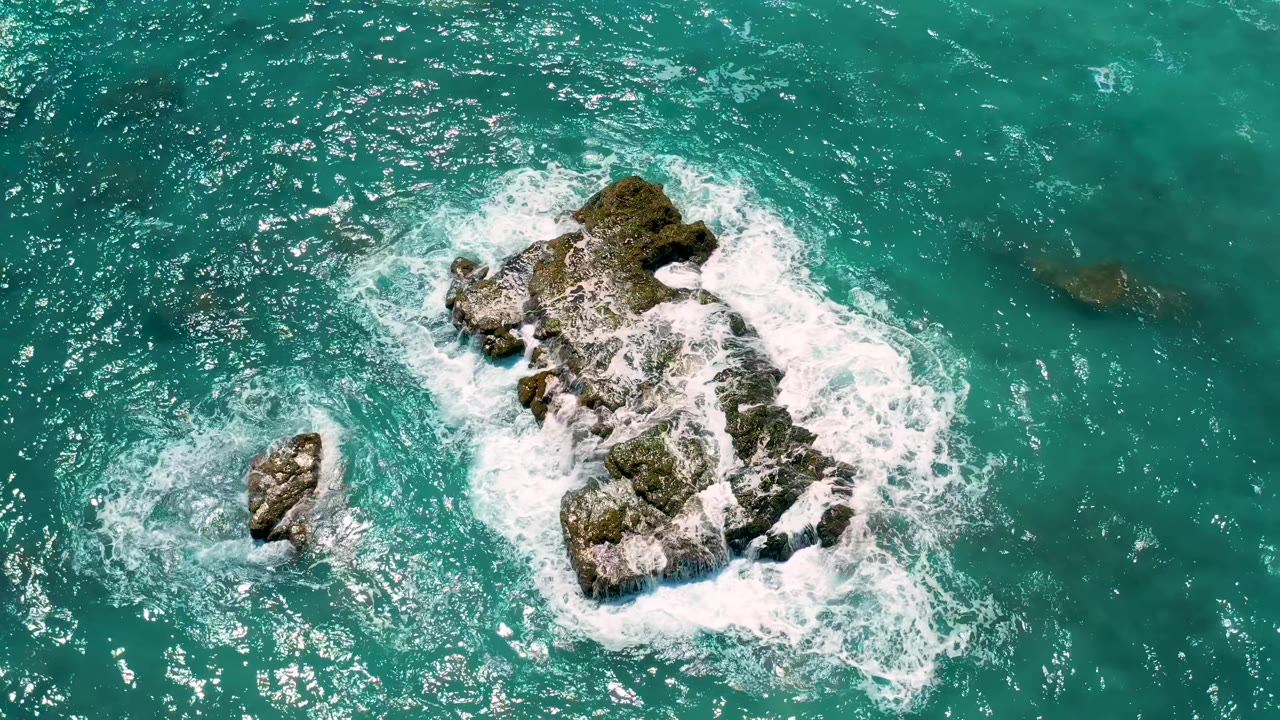 Overhead El Cardoncito frame showing the large rock surrounded by white water, smaller rocks nearby, bright reflected light on the sea, and no visible people.