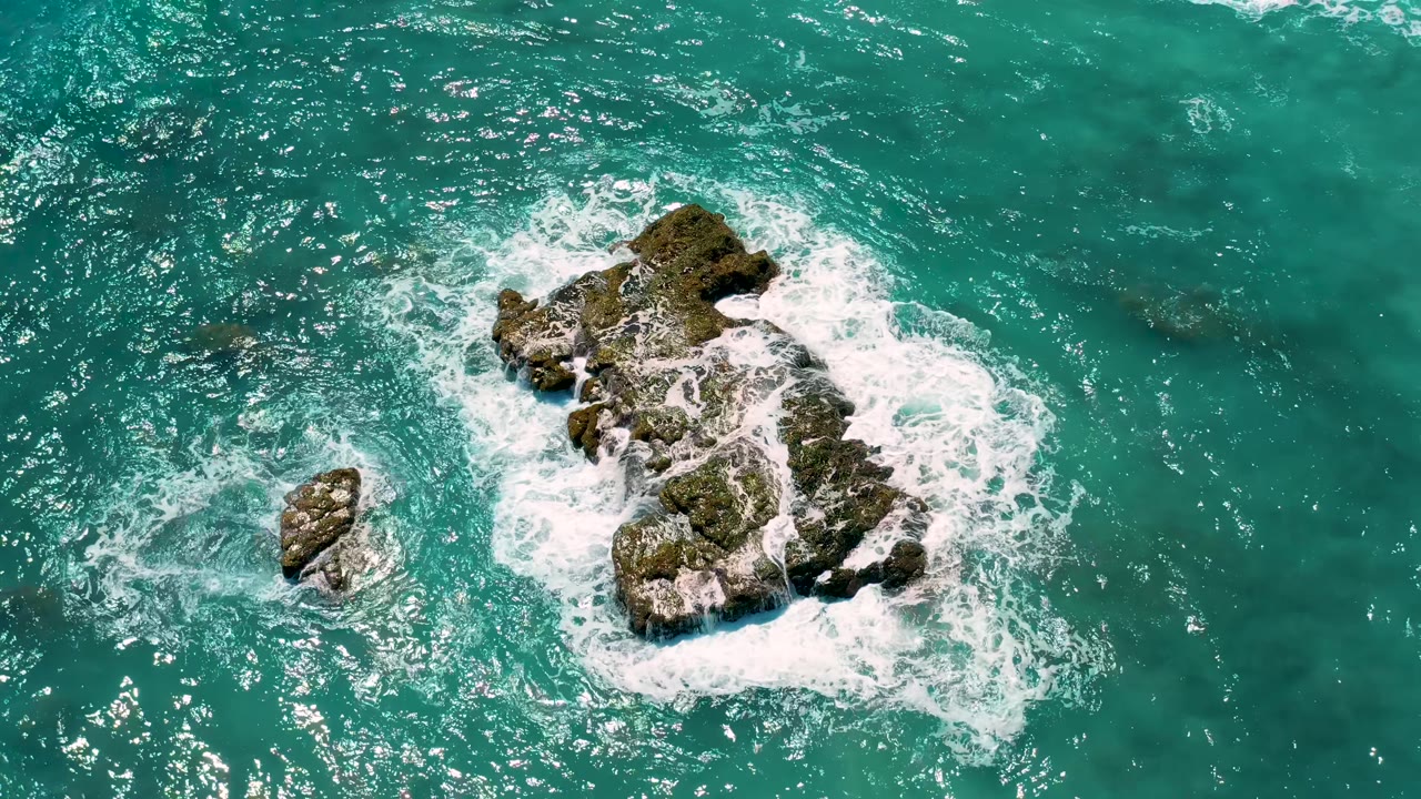 El Cardoncito aerial frame with the main rock lower in the composition, a smaller rock to the left, thin foam trails across turquoise water, and no visible people.