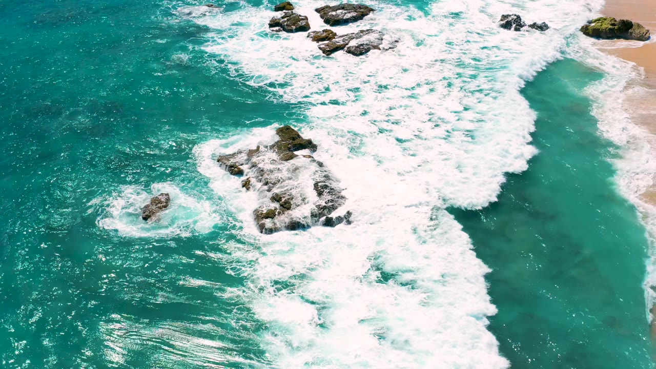 Aerial frame from El Cardoncito with white foam spreading around a large rock formation in blue-green water and no visible people.