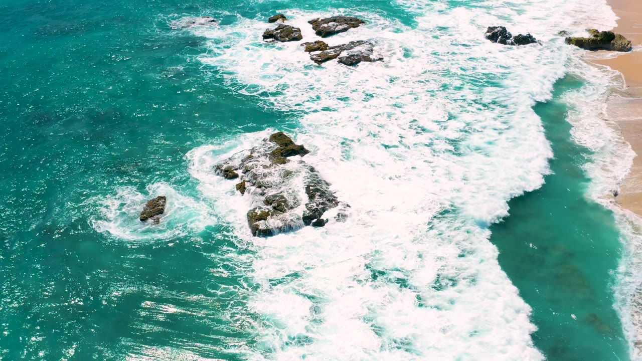 Aerial view of El Cardoncito surf as dense foam covers rocks near the sandy shoreline, with no visible people.