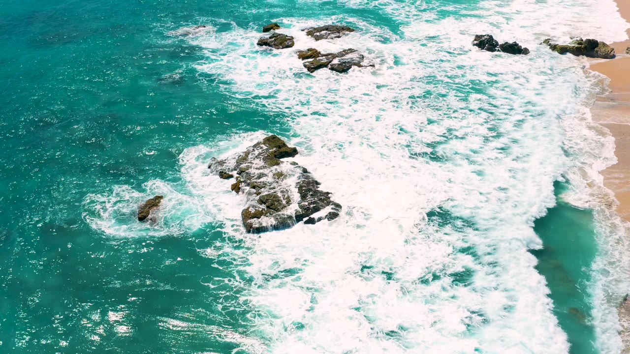 El Cardoncito aerial frame with a central dark rock cluster surrounded by turquoise water and bright white surf, with no visible people.