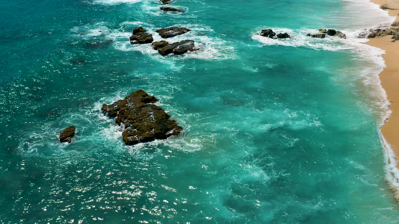 Aerial view at El Cardoncito, Baja California Sur showing turquoise water, dark rock outcrops, white foam, a narrow sand edge on the right, and no visible people.