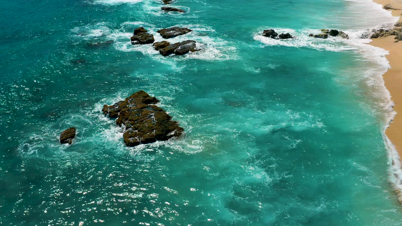 High-angle frame from El Cardoncito with foamy waves wrapping dark rocks in clear teal water, pale sand along the right edge, and no visible people.