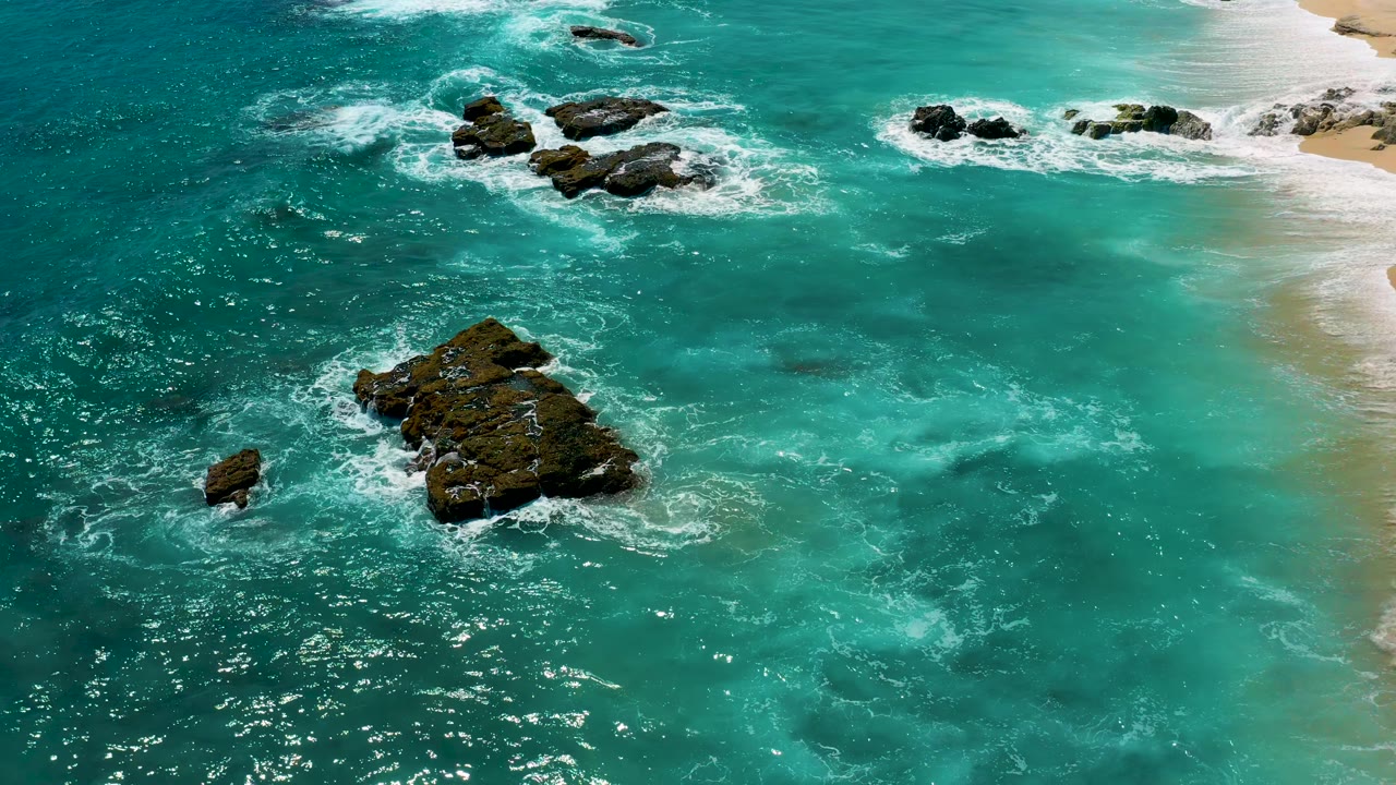 Aerial El Cardoncito frame showing a central dark rock cluster, swirling white surf, deeper blue water to the left, pale beach at the upper right, and no visible people.
