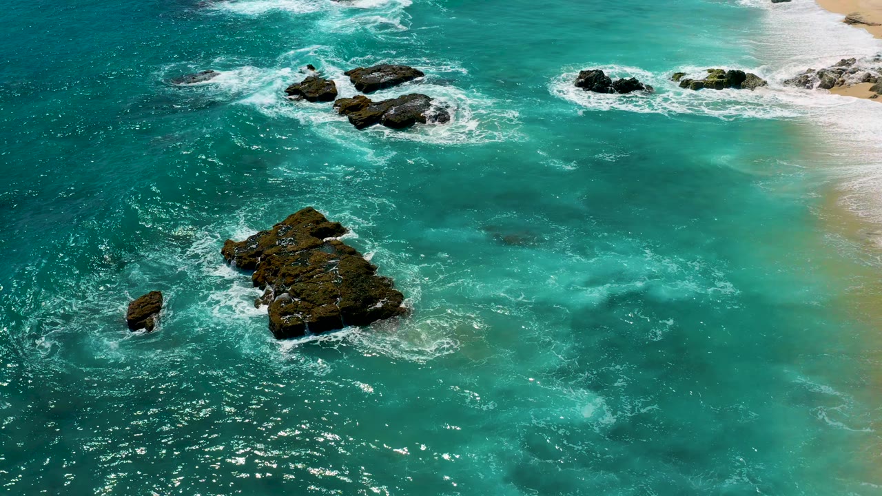 Aerial view over El Cardoncito water with a large lower rock, smaller left rock, foam rings around upper rocks, sandy shoreline on the right, and no visible people.