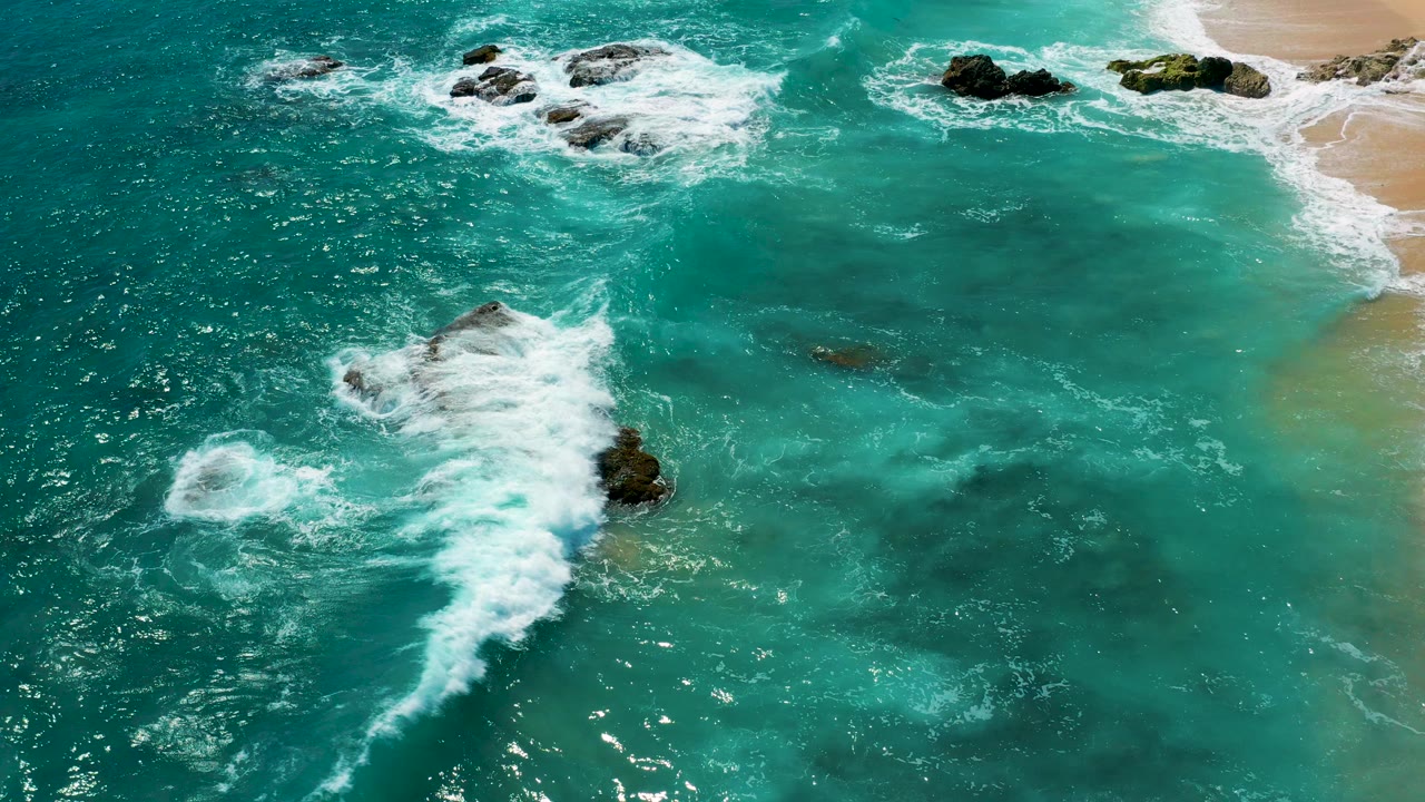Overhead view of turquoise water and white surf spreading around dark rocks near the sandy edge of El Cardoncito, Baja California Sur, with no visible people.