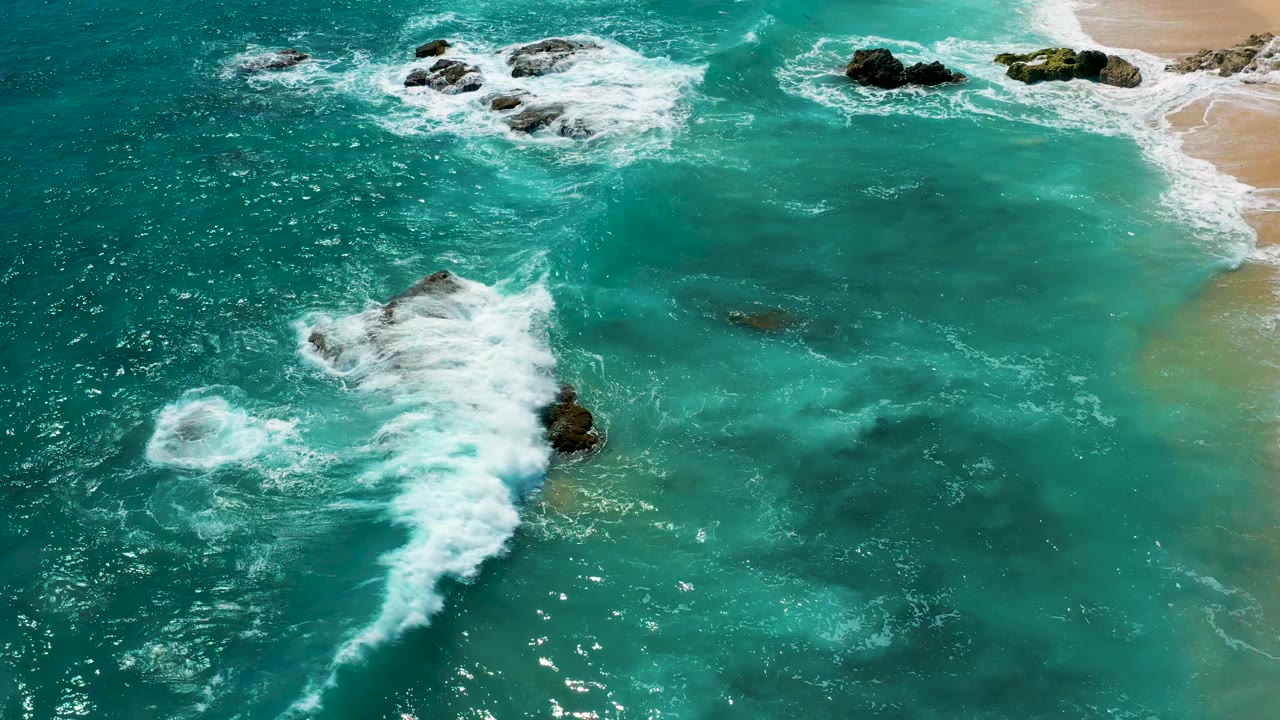 A white wave plume pushes across bright green water toward dark rocks and a pale sand shoreline at El Cardoncito, Baja California Sur, with no visible people.