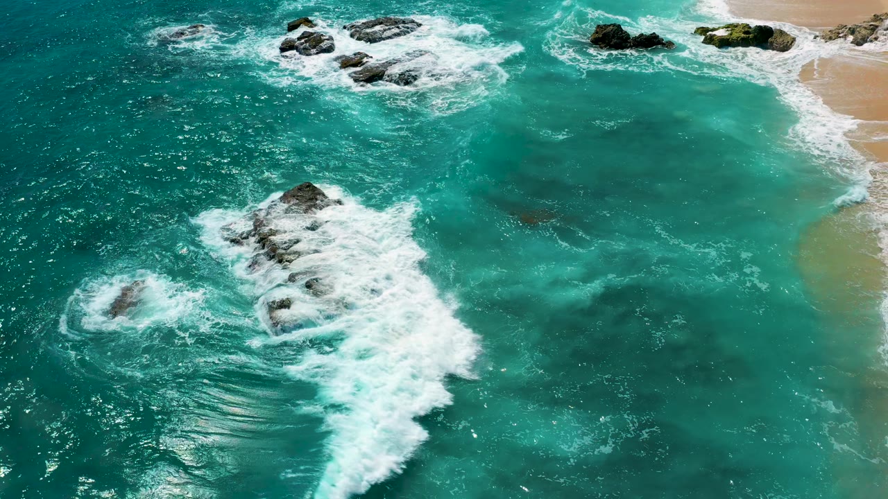 White foam curls around a central dark rock formation while translucent green water and the beach edge remain visible at El Cardoncito, Baja California Sur, with no visible people.