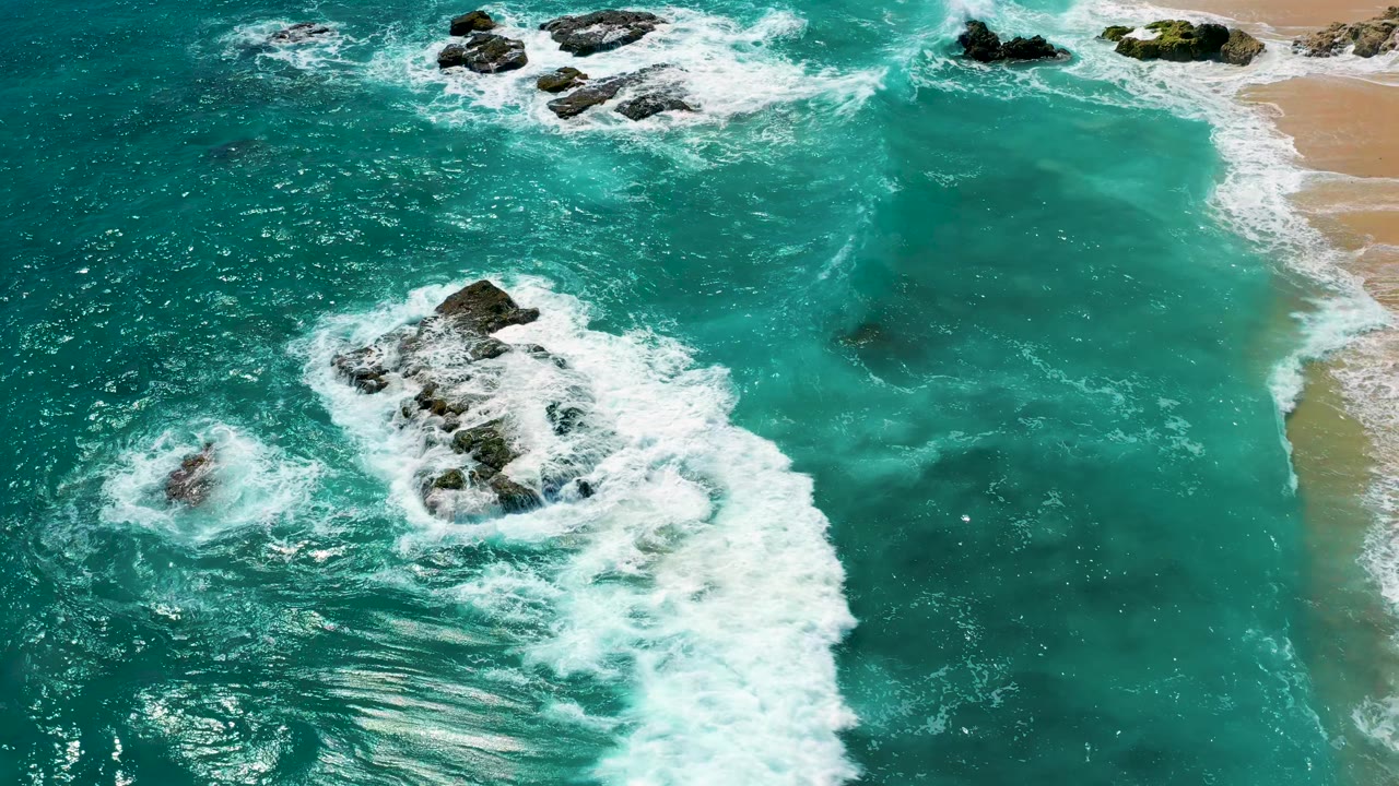 From above, white surf surrounds clustered dark rocks in green water, with the sandy shoreline running along the right side at El Cardoncito, Baja California Sur, and no visible people.