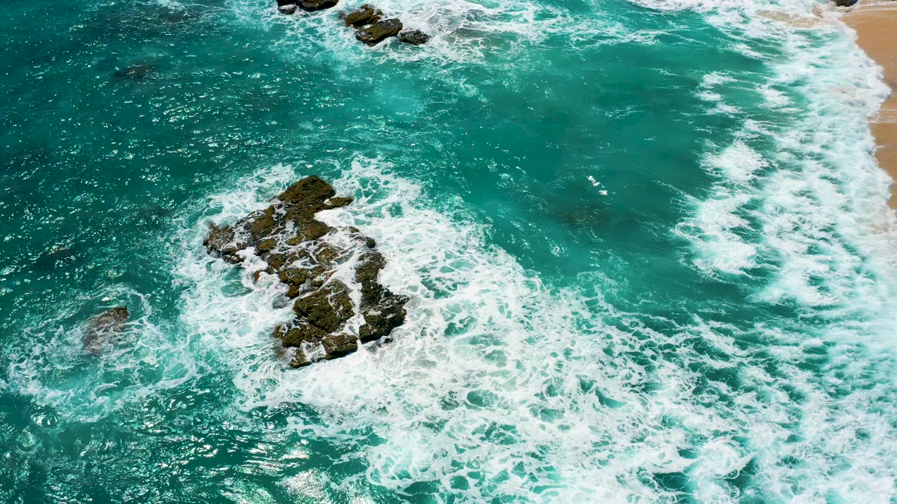 Overhead frame at El Cardoncito, Baja California Sur with foamy surf curling around the central rock formation and a pale sand edge near the upper right, with no visible people.