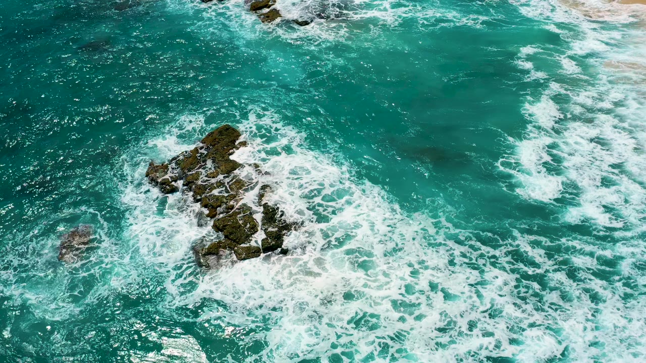 Overhead El Cardoncito frame showing the rock cluster slightly higher in the composition as white water spreads across the surrounding blue green sea, with no visible people.