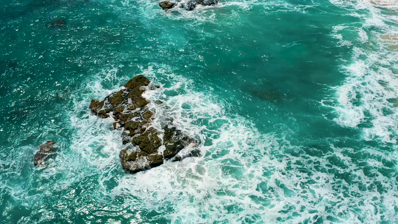 Aerial view at El Cardoncito, Baja California Sur where the main rocks sit left of center and a band of breaking white water runs along the right side, with no visible people.