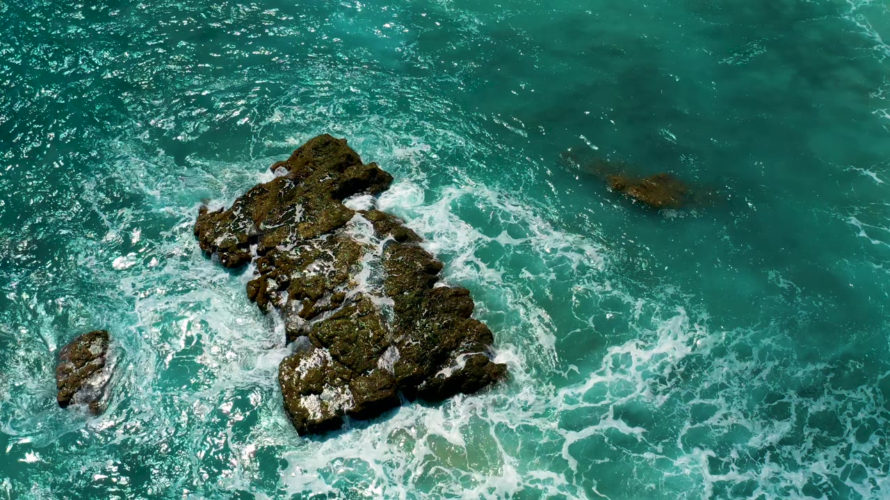 Aerial view at El Cardoncito with the main rock slightly right of center, foamy water below it, a smaller rock at lower left, and no visible people.