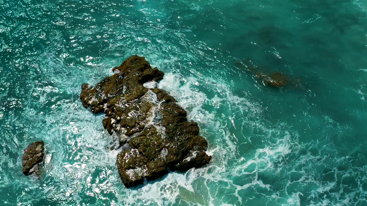 Overhead El Cardoncito frame with bright sun sparkle on the upper water, a dark rock mass on the right, white foam around its base, and no visible people.
