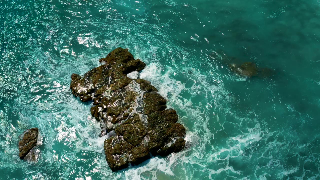 Aerial frame from El Cardoncito showing the rock formation larger on the left side, whitewater trailing into clear turquoise water, and no visible people.