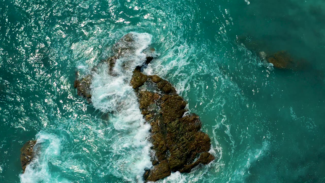Top-down view of turquoise water and white foam breaking around mossy rock formations in El Cardoncito, Baja California Sur, with no visible people.