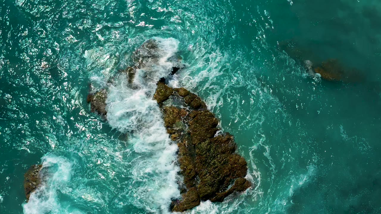 Aerial frame with a long mossy rock angled through turquoise water as white foam spreads across its upper edge in El Cardoncito, with no visible people.