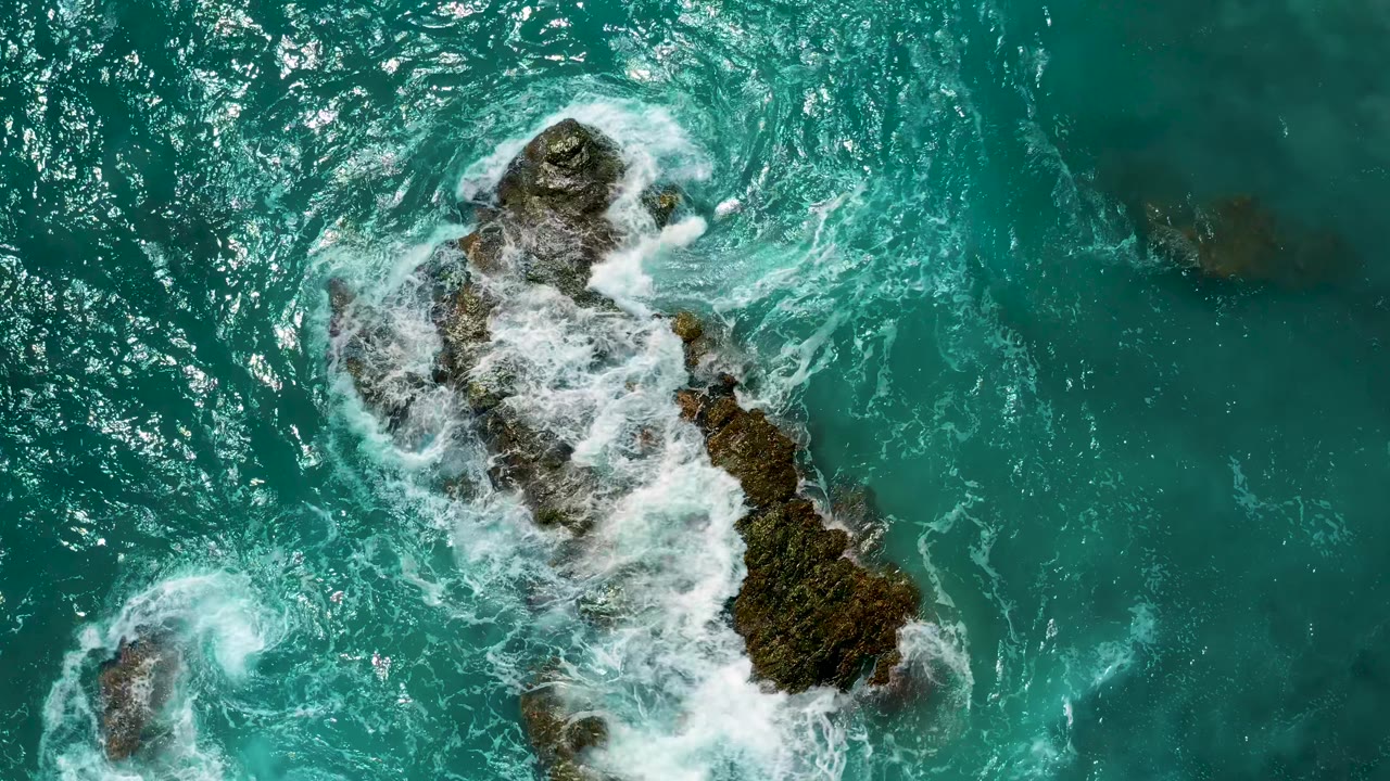 White water rings the upper rock and trails along a larger mossy outcrop below in clear blue ocean at El Cardoncito, with no visible people.