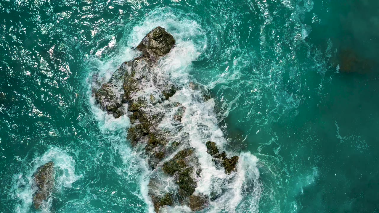 Aerial view of a jagged mossy rock formation partly covered by swirling white foam in turquoise water at El Cardoncito, with no visible people.