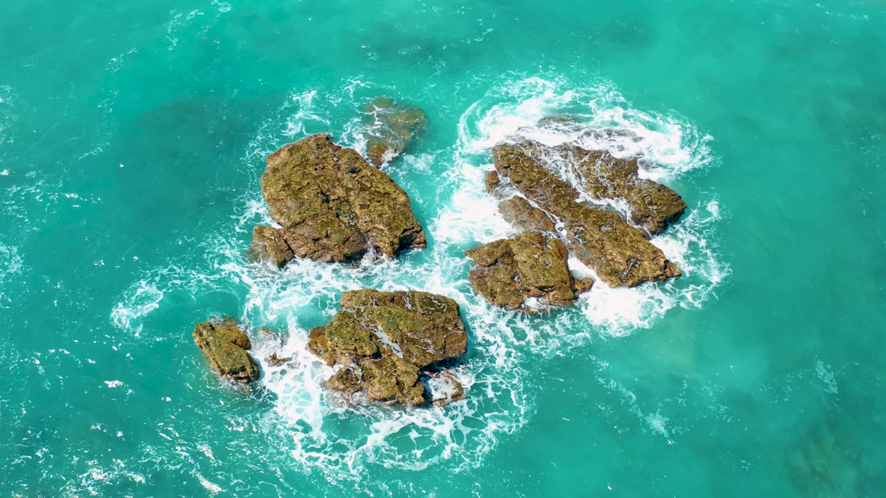 Aerial frame over El Cardoncito where blue-green water fills the view and white waves wrap around the right-side rocks, with no visible people.