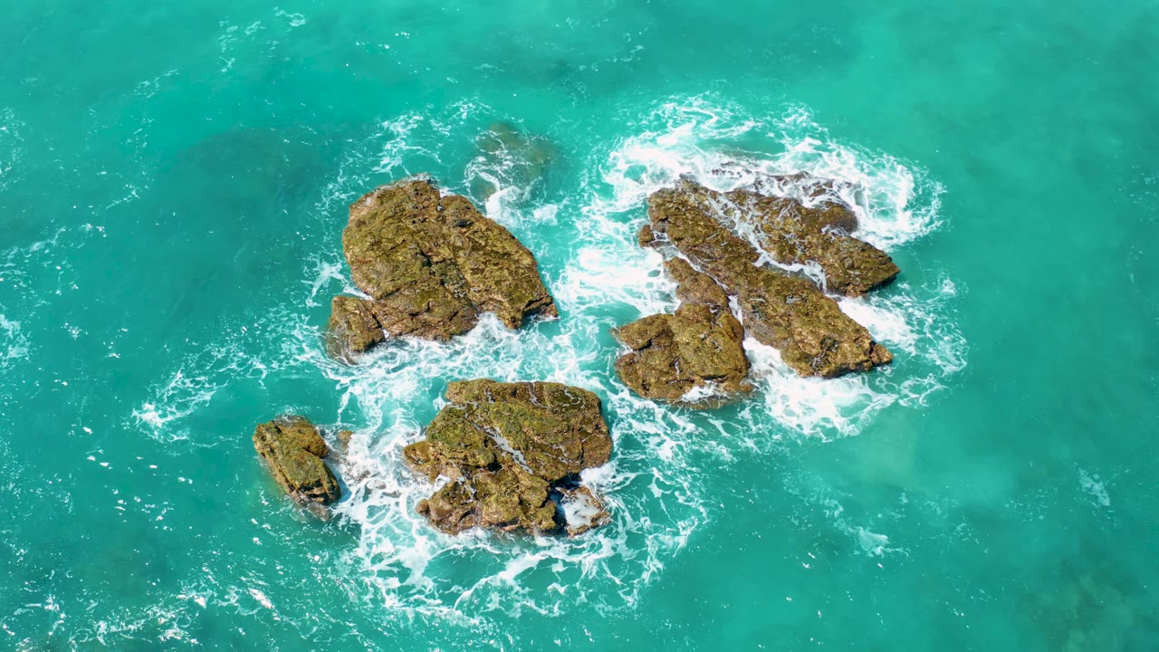 Aerial view of the El Cardoncito rock group with fresh white foam around the lower rocks and turquoise water across the full frame, with no visible people.