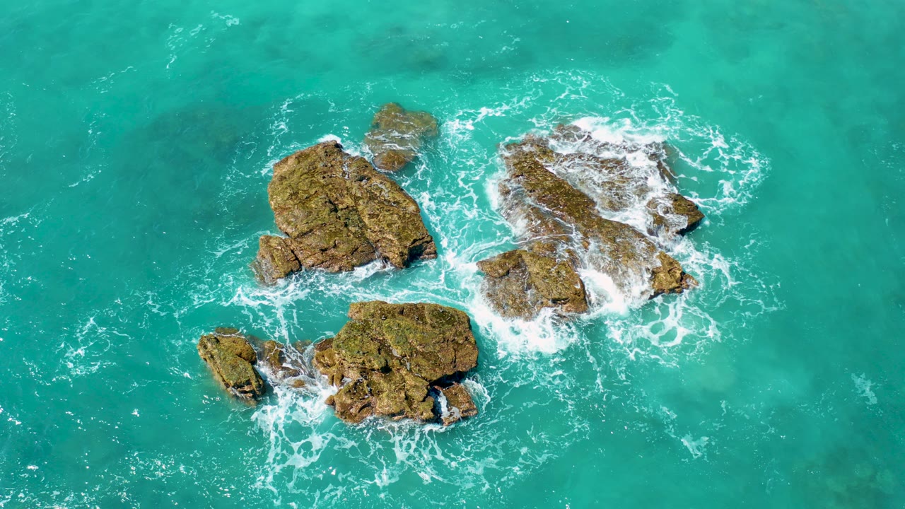 Overhead view at El Cardoncito, Baja California Sur, showing clustered brown rocks in turquoise ocean as white foam gathers around the edges, with no visible people.