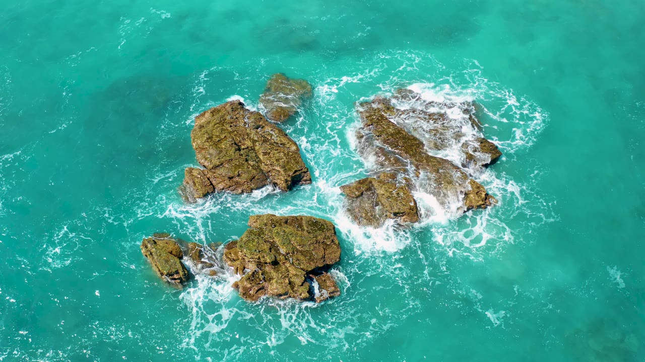 Aerial frame from El Cardoncito, Baja California Sur, with foamy water spreading over the upper right rock while smaller rock outcrops sit in clear turquoise water, with no visible people.